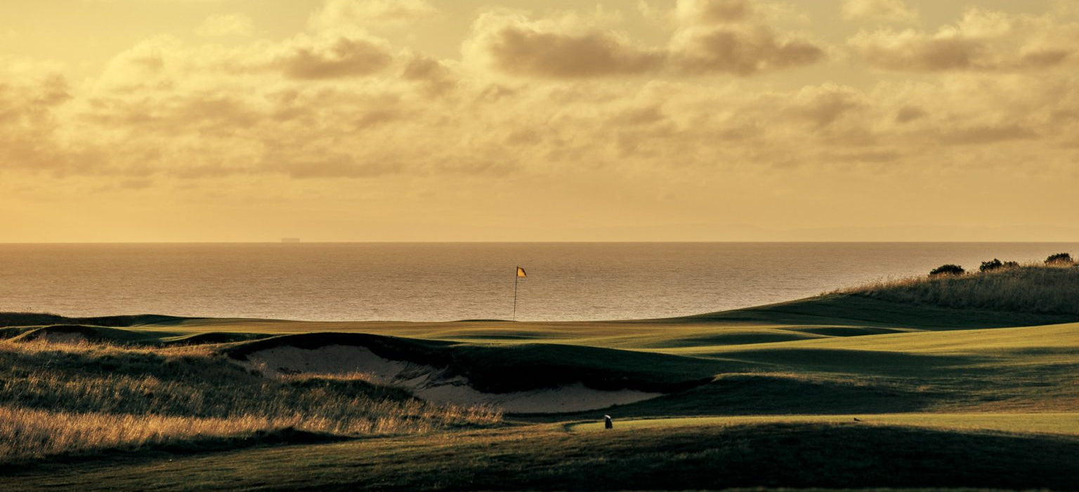A scenic golf course landscape at golden hour with rolling green fairways, sand bunkers, a distant flag, ocean water, and a cloudy sky in warm yellow and brown tones.