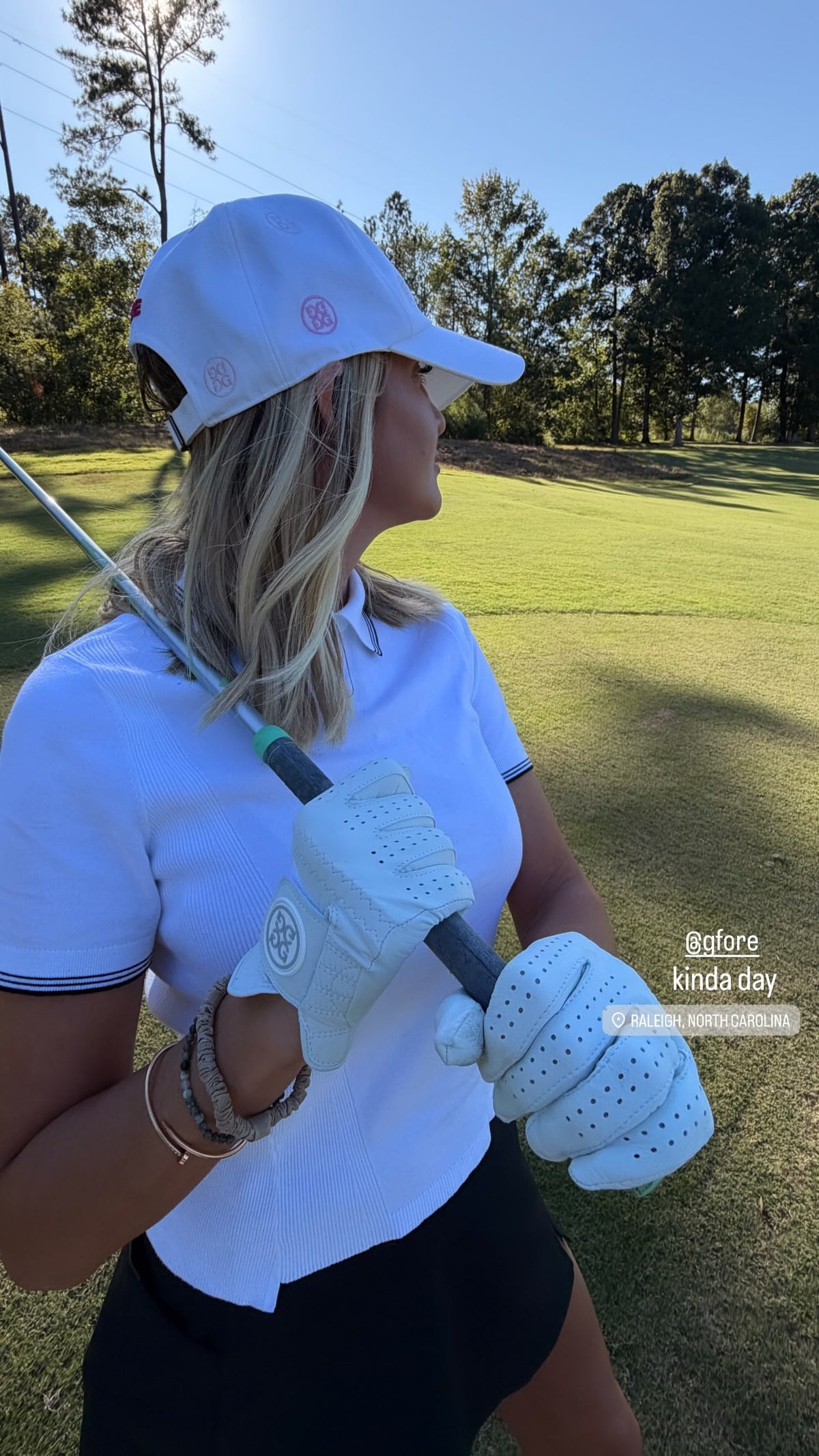 A woman on a golf course wearing a white polo shirt, a white cap, white golf gloves, and a black skirt, holding a golf club. The background shows a green grass field and trees under a blue sky.